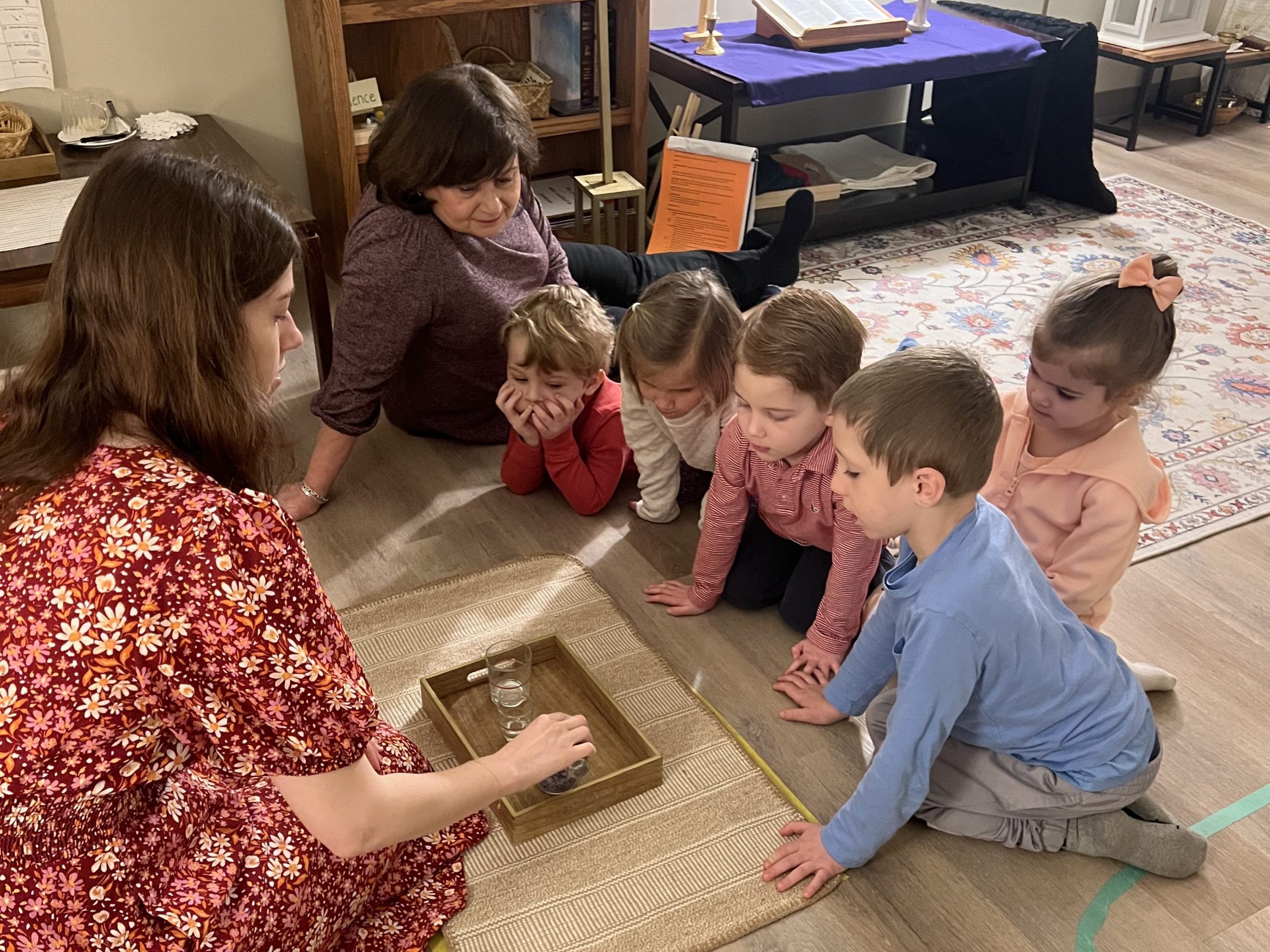 Preschool students kneeling around an adult presenting objects in the Catechesis of the Good Shepherd Atrium.